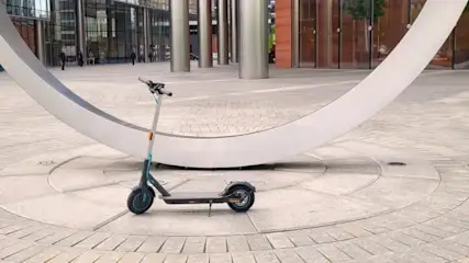 Na placu stoi hulajnoga elektryczna. W tle budynki z kolumnami i biała, łukowa rzeźba. Nawierzchnia placu z kostki, ułożona w okręgi.