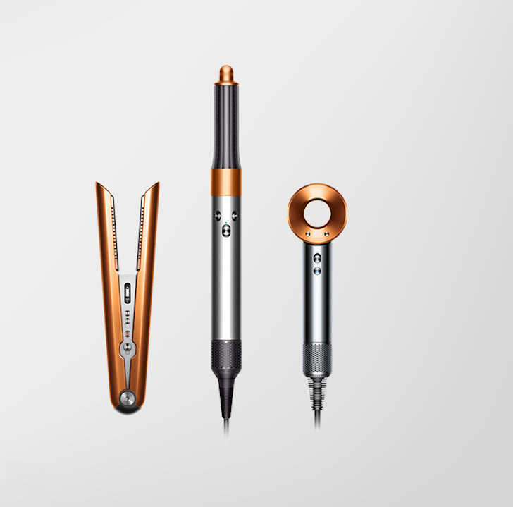 Na jasnoszarym tle leżą prostownica, lokówka i suszarka do włosów w kolorach srebrnym i miedzianym.