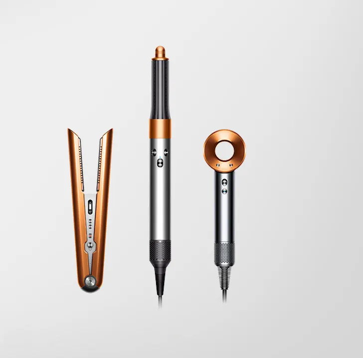 Na jasnoszarym tle leżą prostownica, lokówka i suszarka do włosów w kolorach srebrnym i miedzianym.