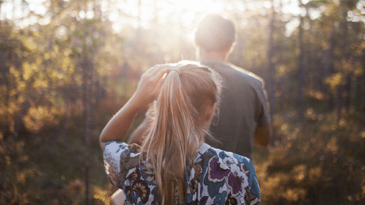 Kobieta w kwiecistej bluzce wiąże włosy w kucyk, obok stoi mężczyzna w szarej koszulce. W tle drzewa i promienie słoneczne.