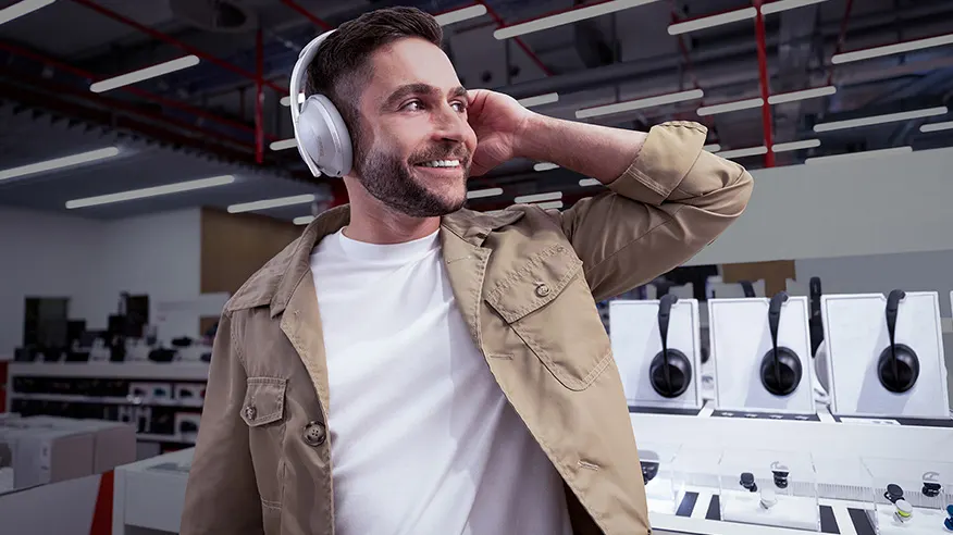 Mężczyzna w białych słuchawkach, w sklepie z elektroniką. Nosi beżową kurtkę i białą koszulkę. Na półkach widoczne słuchawki.