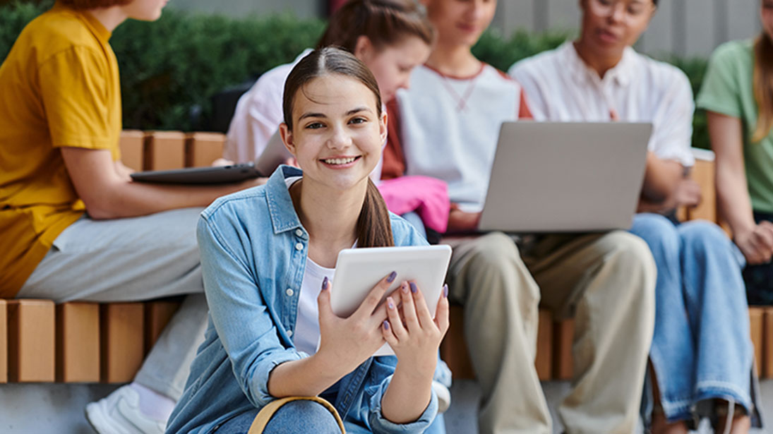 Grupa osób siedzi na ławce; jedna trzyma tablet, inni używają laptopa i tabletu. W tle widać zielone rośliny.