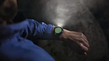 Osoba w niebieskiej koszuli patrzy na zegarek z zielonym ekranem w ciemnym otoczeniu.