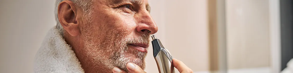 Mężczyzna z brodą przycina włosy w nosie srebrnym trymerem, ubrany w biały ręcznik.