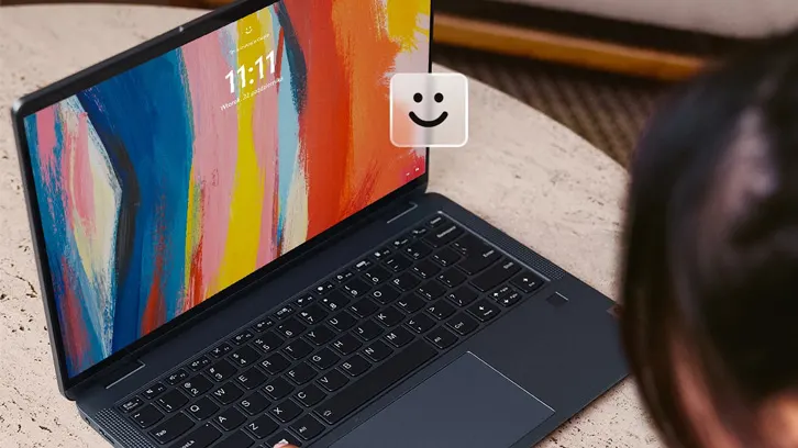 Laptop z kolorowym ekranem pokazuje godzinę 11:11. Na ekranie widoczna ikona uśmiechu. Obok laptopa osoba.