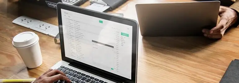 Osoba pisze na laptopie przy biurku. Obok stoi kubek z napojem i listwa zasilająca. W tle drugi laptop i inne przedmioty biurowe.