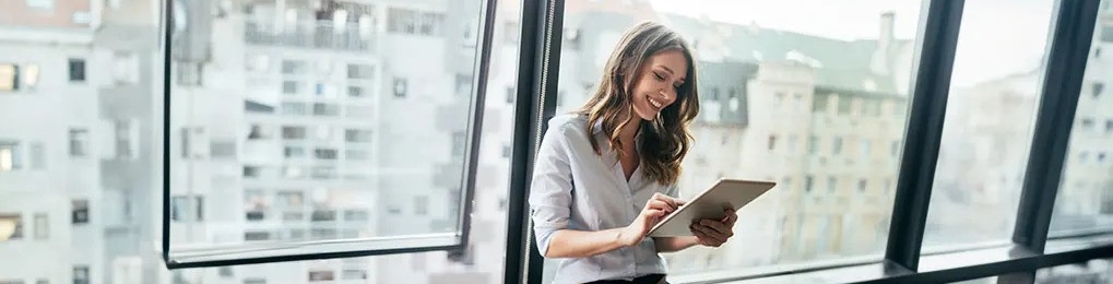 Kobieta używa tabletu blisko okna. Za oknem widać budynki miejskie.