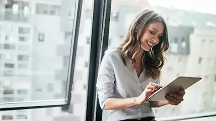 Kobieta używa tabletu blisko okna. Za oknem widać budynki miejskie.