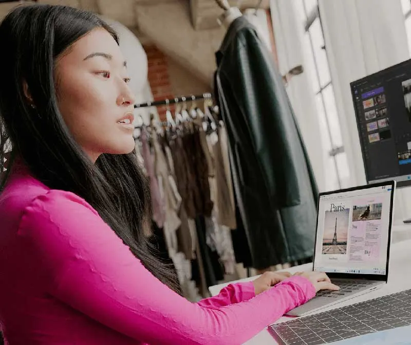 Kobieta w różowej bluzce pracuje na laptopie. W tle widać ubrania na wieszakach i monitor komputera.
