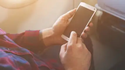 Osoba w koszuli w kratę trzyma telefon komórkowy przy oknie samolotu.