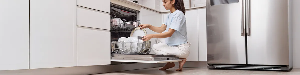 Kobieta obsługuje zmywarkę wypełnioną białymi talerzami w metalowym koszu. Widoczny panel sterowania i wnętrze kuchni.