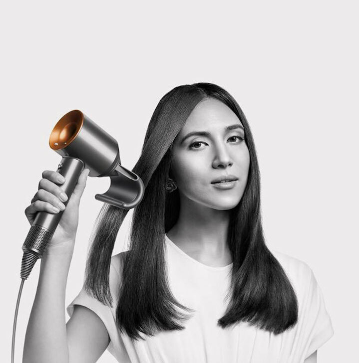 Kobieta stylizuje włosy suszarką z metaliczną, złotą końcówką na jasnym tle. Ma na sobie białą koszulkę i kolczyki.