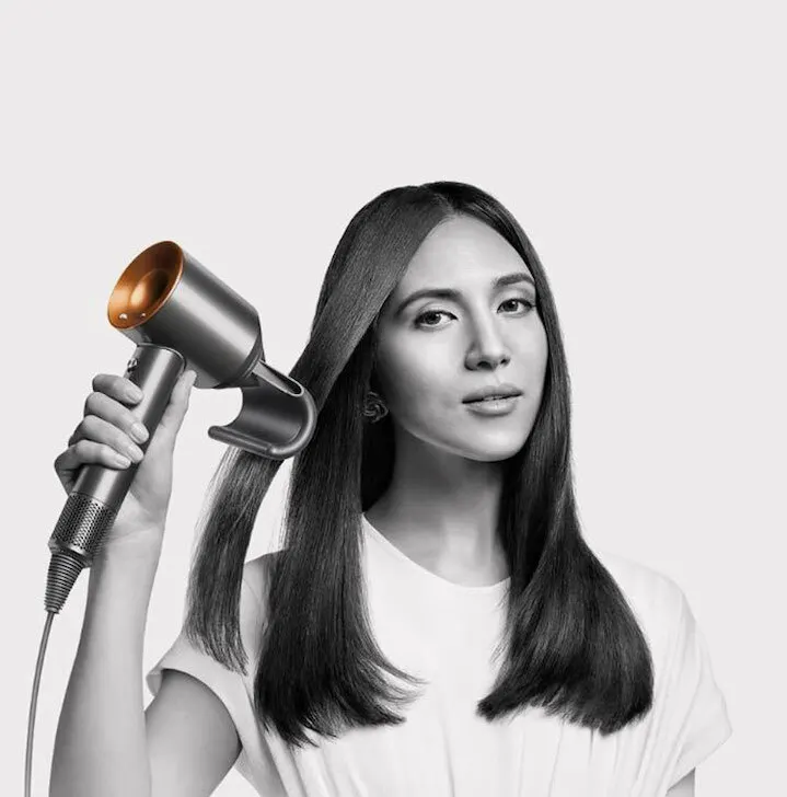 Kobieta stylizuje włosy suszarką z metaliczną, złotą końcówką na jasnym tle. Ma na sobie białą koszulkę i kolczyki.