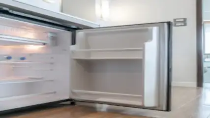 Otwarta lodówka z półkami w jasnym pokoju. W tle widoczne okno i jasne płytki podłogowe.