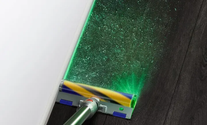 Szczotka odkurzacza z zielonym światłem oświetla drobinki kurzu na ciemnej podłodze. Obok biała ściana.