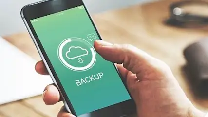 Osoba trzyma telefon z zielonym ekranem, na którym widnieje ikona chmury i napis "Backup". W tle leżą okulary i notatnik.