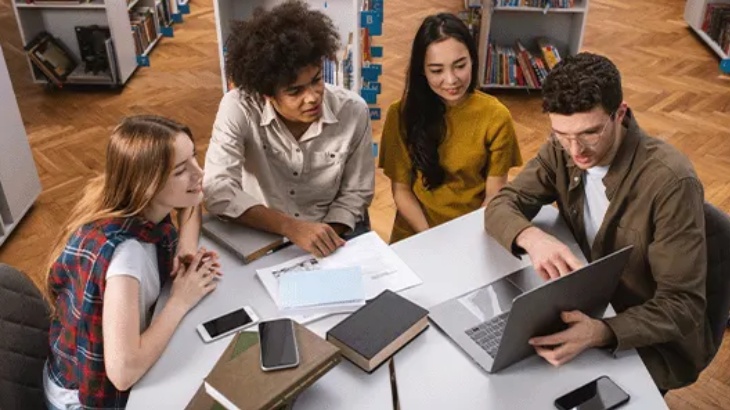 Cztery osoby siedzą przy stole w bibliotece, używając laptopa, książek i telefonów komórkowych. Półki z książkami widoczne w tle.