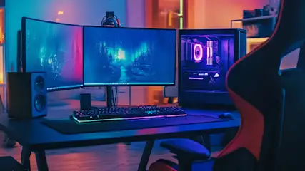 Pokój z biurkiem, komputerem, monitorami i krzesłem do gier, oświetlony niebieskim i pomarańczowym światłem. Na półkach książki i roślina.
