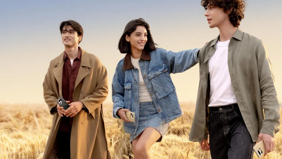 Trzech młodych ludzi idzie przez pole dojrzałego zboża przy łagodnym, złotym świetle. Po lewej stronie stoi mężczyzna w brązowym płaszczu i bordowej koszuli, trzymający w dłoni ciemny smartfon. Pośrodku znajduje się kobieta w dżinsowej kurtce i spódniczce, uśmiechająca się i trzymająca w dłoni złoty smartfon. Po prawej stronie idzie mężczyzna w oliwkowej koszuli i białej koszulce, również trzymający w dłoni złoty smartfon realme 16 pro+ 5g.