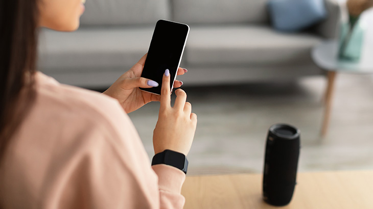 Kobieta trzyma telefon z czarnym ekranem. Na stole głośnik i smartwatch. W tle sofa w salonie.