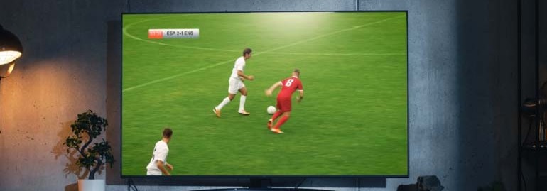 Na ekranie telewizora widać mecz piłki nożnej na zielonym boisku, gdzie zawodnicy w białych i czerwonych strojach grają w piłkę.