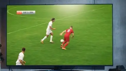 Na ekranie telewizora widać mecz piłki nożnej na zielonym boisku, gdzie zawodnicy w białych i czerwonych strojach grają w piłkę.