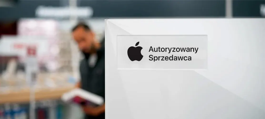 Biały szyld z czarnym logo Apple i napisem "Autoryzowany Sprzedawca". W tle rozmazana postać mężczyzny czytającego broszurę.