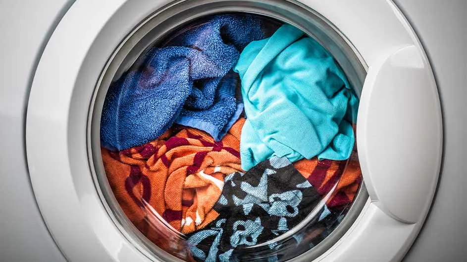 Pralka automatyczna z otwartymi drzwiami, wypełniona kolorowymi ubraniami. Widoczne tkaniny w odcieniach niebieskiego, pomarańczowego i czarnego.