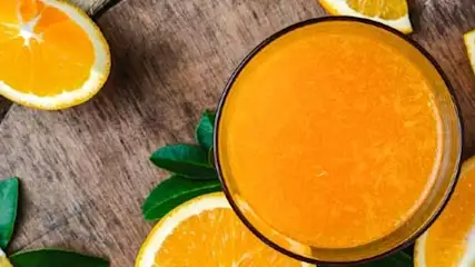 Na drewnianym tle szklanka soku pomarańczowego otoczona kawałkami pomarańczy i zielonymi liśćmi.