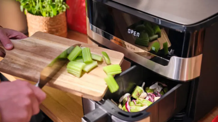 Osoba wrzuca seler z deski do kosza na śmieci w nowoczesnym, czarno-srebrnym urządzeniu kuchennym. Z boku stoi roślina w doniczce.