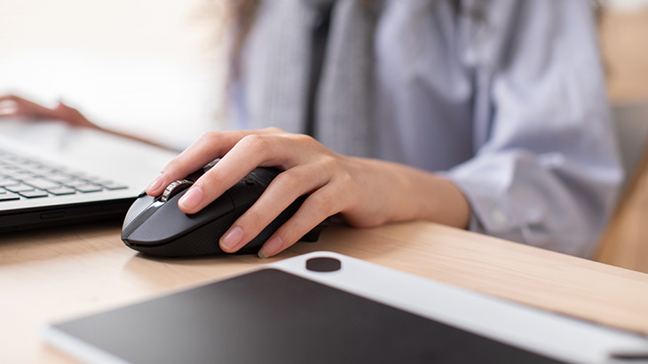 Na stole leży laptop, tablet graficzny i czarna myszka komputerowa, z której korzysta osoba w błękitnej koszuli.