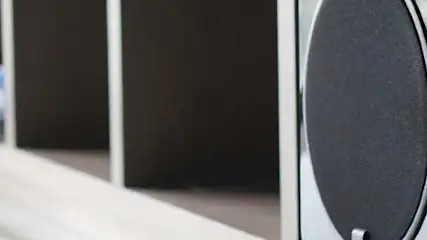 Widok z bliska na głośnik z okrągłą, czarną membraną i białą obudową, ustawiony obok pionowych, ciemnych paneli.