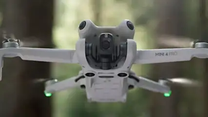 Biały dron z zielonymi światłami leci na tle lasu.