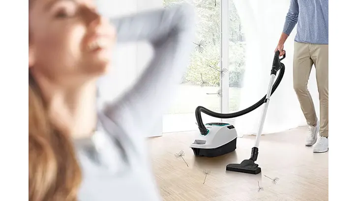Na pierwszym planie kobieta. W tle, mężczyzna odkurza podłogę odkurzaczem w kolorze białym i czarnym. Przez okno widać zieleń.