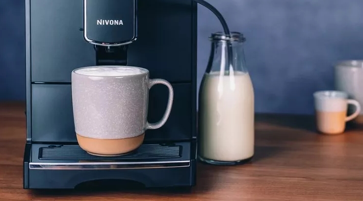 Ekspres do kawy Nivona z kubkiem i spienionym mlekiem obok szklanej butelki z mlekiem na drewnianym stole, w tle niebieska ściana.