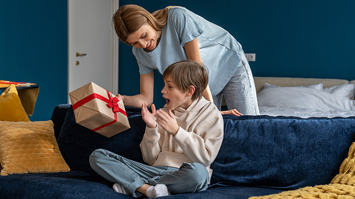 Kobieta daje chłopcu prezent na łóżku z niebieskim zagłówkiem. Chłopiec siedzi i jest zaskoczony. W tle widoczne białe poduszki.