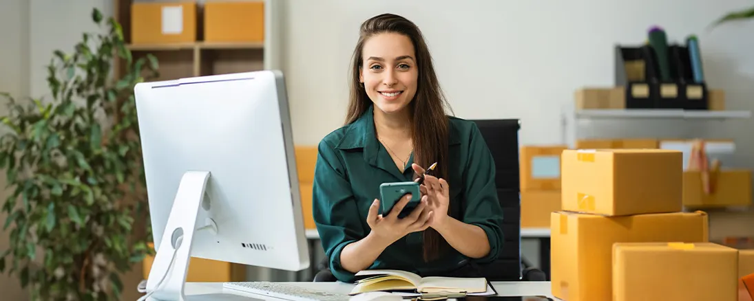 Kobieta siedzi przy biurku z komputerem i paczkami. W rękach trzyma telefon i długopis. Za nią regał i roślina.