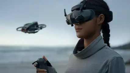 Kobieta w okularach VR steruje dronem na plaży. W tle widać morze i wzgórza.