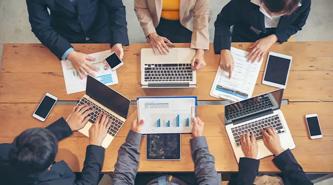 Na drewnianym stole leżą laptopy, tablety, telefony i dokumenty z wykresami. Wokół siedzą osoby w strojach biznesowych.