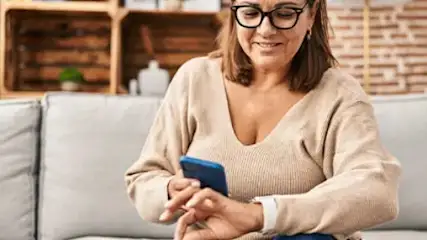 Kobieta w okularach siedzi na szarej kanapie i używa niebieskiego smartfona. Za nią widać ceglaną ścianę i drewniane półki.