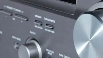 Szary panel z metalowymi pokrętłami i przyciskami do sterowania urządzeniem audio. Widoczne napisy funkcyjne.