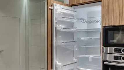 Otwarta lodówka obok piekarnika i mikrofalówki w zabudowie. Na ścianie białe drzwi.