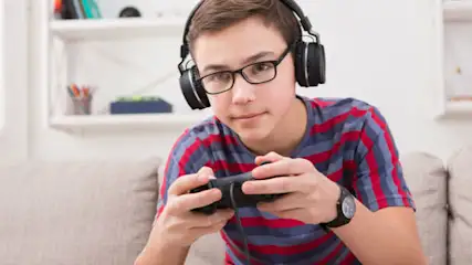 Chłopak w okularach i słuchawkach gra na konsoli, ubrany w koszulkę w czerwono-szare paski, siedzi na beżowej kanapie. Za nim białe półki.