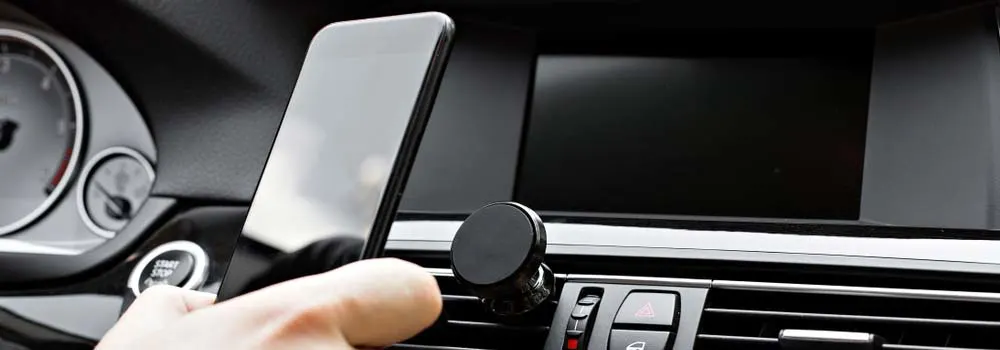 Wnętrze samochodu z czarnym ekranem i uchwytem na telefon w kratce wentylacyjnej. Ręka trzyma smartfon.