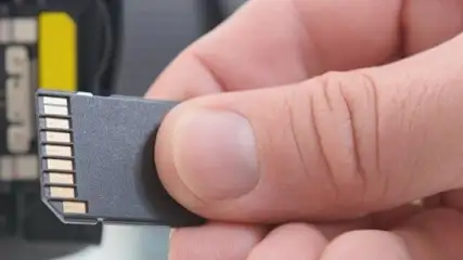 Dłoń trzyma czarną kartę pamięci z metalowymi stykami, w tle czarne urządzenie z żółtym elementem.