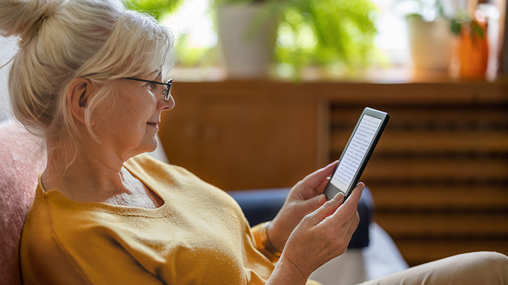 Kobieta w okularach trzyma czytnik e-booków, siedząc na kanapie. W tle widać rośliny doniczkowe i drewniane meble.