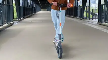 Osoba jedzie na hulajnodze po kładce dla pieszych z czarnymi barierkami. W tle widoczny budynek.