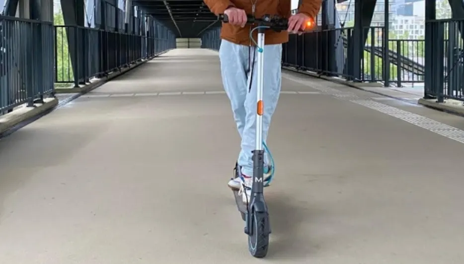 Osoba jedzie na hulajnodze po kładce dla pieszych z czarnymi barierkami. W tle widoczny budynek.