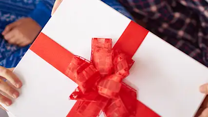 Prezenty na szarym tle. Białe pudełko z czerwoną wstążką trzyma dziecko w niebieskiej piżamie i osoba w koszuli w kratę.