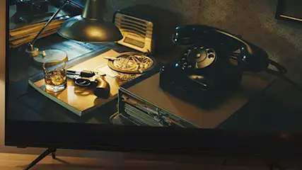 Na ekranie telewizora widoczny jest stary telefon, popielniczka, rewolwer i szklanka alkoholu. Obok stoi lampa podłogowa.